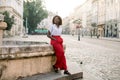 Outdoor full length portrait of young pretty African business lady, wearing red pants and striped shirt, resting after Royalty Free Stock Photo