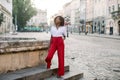 Outdoor full length portrait of young pretty African business lady, wearing red pants and striped shirt, resting after Royalty Free Stock Photo