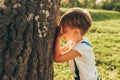 Outdoor image of cute little boy covering his eyes with hands, playing hide and seek standing next a big tree on sunlight and Royalty Free Stock Photo