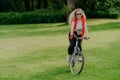 Outdoor image of pretty cheerful young woman rides bike, wears sunglasses, casual wear, poses on green lawn, spends free time in Royalty Free Stock Photo