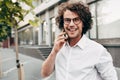 Outdoor image of a young man with curly hair smiling while talking with his friend on a mobile phone. A happy curly male wearing a Royalty Free Stock Photo
