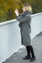 Outdoor lifestyle photo of young beautiful blonde woman in fall autumn park taking photos on smartphone cozy scarf grey vintage co Royalty Free Stock Photo