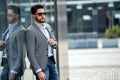 Outdoor photo of handsome elegant man wearing fashionable clothes and sunglasses, posing on the wall with reflections. City stale Royalty Free Stock Photo