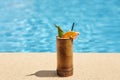 Outdoor picture of wooden cup with cocktail being near swimming pool, green and piece of orange being inside cocktail, black straw