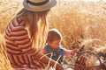 Outdoor picture of young fair haired mother sitting with her little daughter at wheat field, having small picnic together, mum