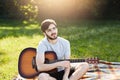 Outdoor portrait of attractive young man with beard dressed casually while resting on green grass with guitar, having pensive expr Royalty Free Stock Photo