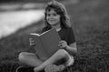 Outdoor portrait of a cute little kid reading a book in summer park. Blond kid boy sitting on grass and reading book Royalty Free Stock Photo