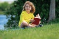 Outdoor portrait of a cute little kid reading a book in summer park. Blond kid boy sitting on grass and reading book Royalty Free Stock Photo