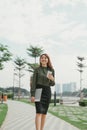 Outdoor portrait of dreamy Asian girl walking In Park With clipboard and coffee in hands