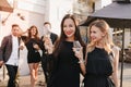 Outdoor portrait of enthusiastic brunette woman in black dress with long sleeves posing with smile near blonde sister