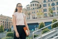 Outdoor portrait of female student 16, 17 years old. Girl in glasses, with backpack, textbooks. City background