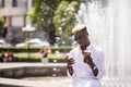 Portrait of handsome afro american young man listening music from phone drinking coffee to go on summer street in front of fountai Royalty Free Stock Photo