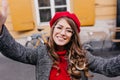 Outdoor portrait of laughing romantic lady in glasses making selfie in front of yellow building. Curly girl in red beret Royalty Free Stock Photo