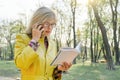 Outdoor portrait of mature business woman with glasses reading book talking on the phone. Female blonde in yellow raincoat, Royalty Free Stock Photo