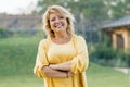 Outdoor portrait of positive confident mature woman. Smiling female blonde in a yellow dress with arms crossed near the house Royalty Free Stock Photo