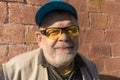 Portrait of smiling Caucasian senior man wearing blue cap and yellow sunglasses while standing against fulvous brick wall Royalty Free Stock Photo