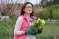 Outdoor portrait of smiling middle-aged woman in garden gloves with flowers for planting, spring flowering garden background, copy