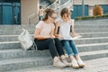 Outdoor portrait of two young beautiful girls students with backpacks, books. Girls sitting on the steps, talking, looking into a Royalty Free Stock Photo