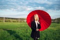 Outdoor portrait of young blonde girl holding red umbrella on background of green field and blue cloudy sky. Woman wearing black. Royalty Free Stock Photo