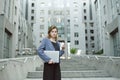 Outdoor portrait of young female enterpreneur standing on stairs of office building holding laptop and coffee. Attractive