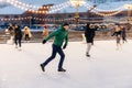 Outdoor shot of attractive man with beard, wears warm winter clothes, practices going skating on ice skate ring decorated with li