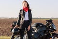 Outdoor shot of attractive woman driver with dark hair stands near black fast motobike, holds helmet, wears leather jacket and red