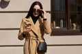 Outdoor shot of attractive young woman wearing coat in sunny city street and drinking take away coffee in paper cup, keeps hand on Royalty Free Stock Photo