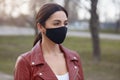 Outdoor shot of beautiful brunette woman wearing leather jacket and black protective mask, looking in distance, protecting herself
