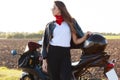 Outdoor shot of confident risky young woman standing near her motorbike, putting hand on helmet, wearing red bandana, white shirt Royalty Free Stock Photo