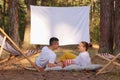 Outdoor shot of couple lying in the forest with overhead projector with white blank screen, eating popcorn, talking and enjoying