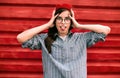 Outdoor shot of funny brunette young woman with hands on head, dressed in casual blue shirt, red beret hat and glasses, making Royalty Free Stock Photo