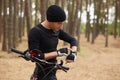 Outdoor shot of handsome man using his smart watch, riding bike in forest, wearing black sportwear and cap, looks athis device,