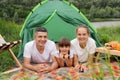 Outdoor shot of happy family, mother, father and cute little daughter with pigtails having picnic near the river, posing near Royalty Free Stock Photo