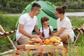 Outdoor shot of happy family sitting near tent on the ground, having picnic together, enjoying delicious fruit and sandwiches, Royalty Free Stock Photo