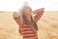 Outdoor shot of happy young woman in striped outfit and sun hat enjoying sun on cereal field, female posing with hands up, looking Royalty Free Stock Photo