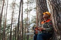 Outdoor shot of logger having rest in open air after cutting trees, sitting near tree with chainsaw and looking at beautiful