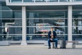 Outdoor shot of male passenger waits for transport on bench, poses at station, looks at watch, suitcase on wheels near, wears