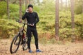 Outdoor shot of man wearing black track suit and sneakers, standing near his mountain bike and speaks phone with beautiful forest Royalty Free Stock Photo