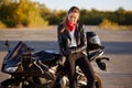 Outdoor shot of pleased female bikes puts on leather gloves, dressed in black clothes, poses on motorbike, prepares for racing or