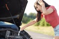 Outdoor shot of puzzled pretty female stares at opened car hood with surprised expression, tries to repair engine by herself, has