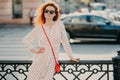 Outdoor shot of satisfied redhead woman keeps one hand on waist, other on street hence, poses over blurred urban setting, wears