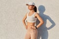 Outdoor shot of slim winsome female wearing sport white top, visor cap and leggins, standing against white wall, holding hands on Royalty Free Stock Photo