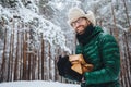 Outdoor shot of smiling glad male with beard and mustache wears spectacles, anork and warm hat, holds firewood, stands against tre