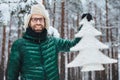 Outdoor shot of smiling unshven middle aged male ears warm cap and green anorak, holds artificial fir tree, enjoy beautiful landsc