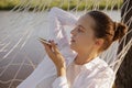 Outdoor shot of young adult woman with bun hairstyle wearing white shirt sitting in hammock on the bank of the river, holding Royalty Free Stock Photo