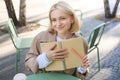 Outdoor shot of young beautiful blond girl with notebook, woman sitting in outdoor cafe, writing in journal, making Royalty Free Stock Photo