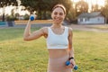 Outdoor shot of young female athlete with dumbbells stretching and warming up in stadium, looking smiling at camera, expressing Royalty Free Stock Photo