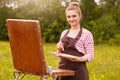 Outdoor shot of young woman artist standing with sketchbook against meadow background, holding brush and palette of colours, Royalty Free Stock Photo