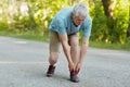 Outdoor view of sporty healthy male pensioner stretches leg, pulled muscle, dressed in sportswear and sneakers, stands on asphalt,