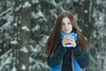 Outdoor winter portrait of freezing young lady holding cup with hot drink during heavy snowfall in conifer forest